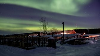 Ein atemberaubendes Nordlicht erhellt den Nachthimmel über einer verschneiten Landschaft mit Holzstrukturen im Vordergrund.