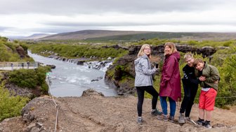 Eine Gruppe von vier Personen steht am Rand eines Wasserfalls, umgeben von grüner Landschaft und einem fließenden Fluss.