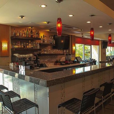 Eine stilvolle Bar mit einem glänzenden Tresen, beleuchtet von roten Pendelleuchten und umgeben von modernen Stühlen.