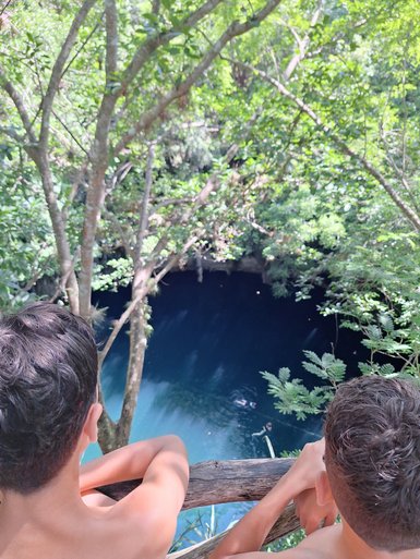 Zwei Jungen lehnen sich über ein Holzgeländer und blicken auf einen tiefblauen Cenote, umgeben von üppigem Grün.