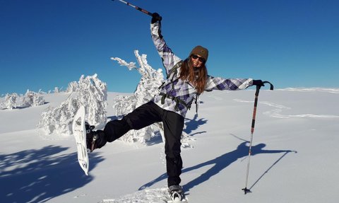 Eine Frau mit Schneeschuhen balanciert fröhlich auf einem schneebedeckten Hang unter einem strahlend blauen Himmel.