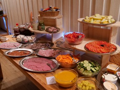 Ein reichhaltiges Frühstücksbuffet mit verschiedenen Wurstsorten, frischem Gemüse und Obst auf Holzplatten angerichtet.