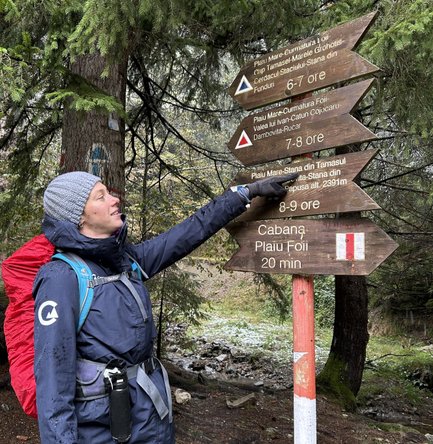 Eine Person in wetterfester Kleidung zeigt auf ein Wegweiser-Schild im Wald, umgeben von grünen Bäumen und einem kleinen Bach.