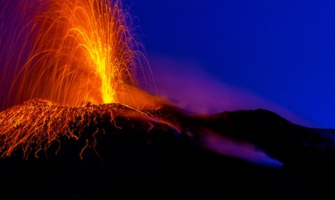 Ein aktiver Vulkan spuckt glühende Lava und Asche in den Nachthimmel, während orangefarbene Funken aufsteigen.