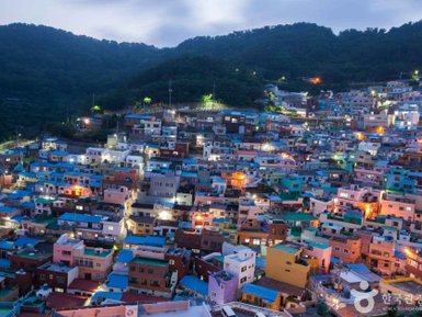 Die bunten Häuser des Gamcheon Culture Village leuchten in der Dämmerung, umgeben von sanften Hügeln und einem klaren Himmel.