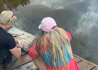 Zwei Personen sitzen am Ufer eines ruhigen Sees, während die Sonne hinter den Bäumen untergeht und das Wasser reflektiert.