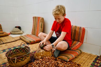 Ein Junge sitzt auf dem Boden und mahlt Argan-Nüsse mit einem Stein in einem hellen Raum mit gestreiften Kissen.