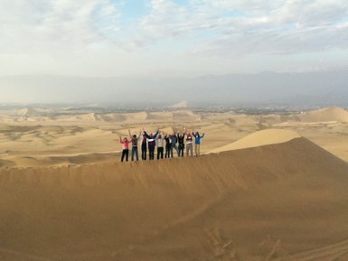 Eine Gruppe von Menschen steht auf einer Sanddüne in der Wüste, alle heben die Arme in die Luft und strahlen Freude aus.