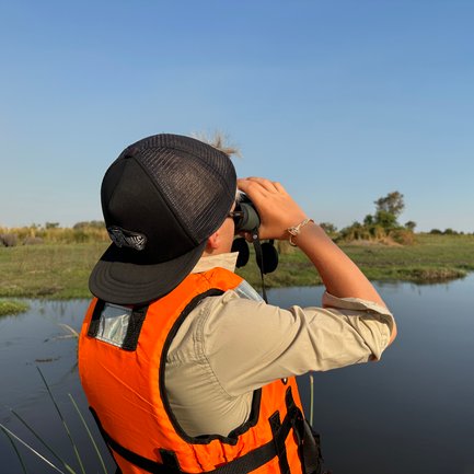 Ein Junge in einer orangefarbenen Weste beobachtet mit einem Fernglas die Landschaft am Wasser, umgeben von üppigem Grün.