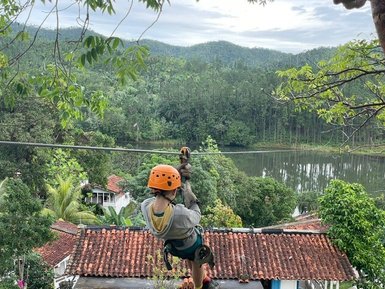 Ein Abenteurer schwingt an einer Zipline über ein malerisches Tal mit üppigem Grün und einem ruhigen See im Hintergrund.