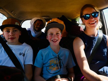 Eine fröhliche Familie sitzt im Auto, während sie durch die Wüste Marokkos fährt, umgeben von Gepäck und Sonnenlicht.