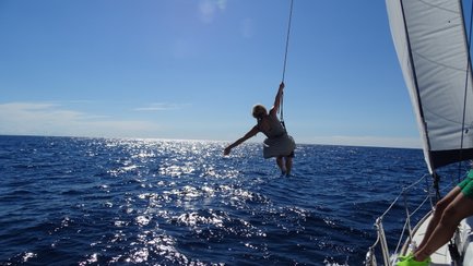 Eine Person schwingt fröhlich über dem glitzernden Wasser, während sie an einem Seil von einem Segelboot hängt.