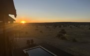 Die Sonne geht über der weiten namibischen Landschaft auf, während ein Pool und Liegen im Vordergrund sichtbar sind.