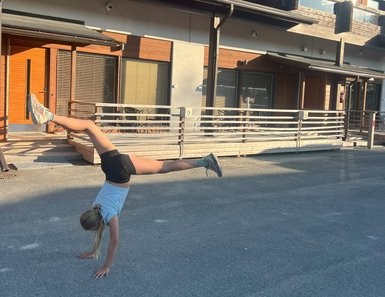 Ein Mädchen macht einen Handstand auf einer Straße vor modernen Holzhäusern, während die Sonne scheint.