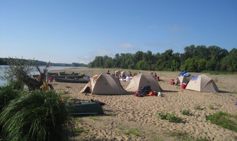 Am Ufer eines Flusses stehen mehrere Zelte auf sandigem Boden, umgeben von Bäumen und Booten, die auf dem Wasser liegen.