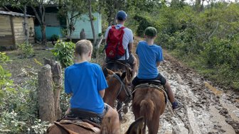 Drei Reiter, darunter zwei Kinder, reiten auf einem schlammigen Weg durch eine grüne Landschaft mit Bäumen.