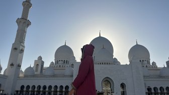 Eine Person in einem roten Gewand steht vor der beeindruckenden Sheikh Zayed Moschee, umgeben von majestätischen Kuppeln.