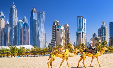 Zwei Kamele mit einem Reiter ziehen durch den Sand, während die beeindruckende Skyline von Dubai im Hintergrund leuchtet.