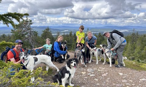 Eine Gruppe von Menschen posiert fröhlich mit mehreren Huskys auf einem Berg, umgeben von einer malerischen Landschaft.