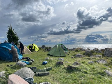 Eine Gruppe von Menschen baut Zelte am Ufer eines Sees auf, umgeben von großen Steinen und einer dramatischen Wolkenlandschaft.