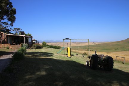 Ein Bauernhof mit einem Spielplatz, einer Schaukel und einem Traktor, umgeben von weitläufigen Feldern und Bergen im Hintergrund.