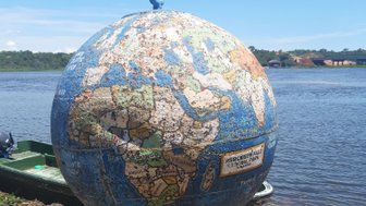 Ein abgenutzter Globus steht am Ufer eines Gewässers, umgeben von Gras und kleinen Steinen, unter einem klaren blauen Himmel.