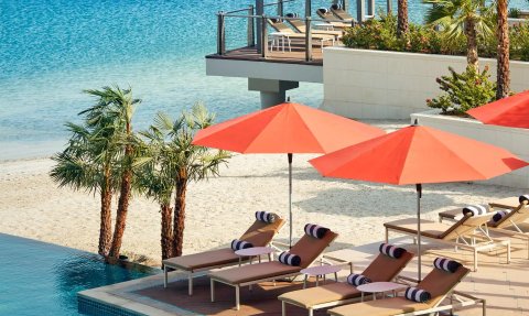 Am Strand stehen mehrere Liegen mit roten Sonnenschirmen, umgeben von Palmen und einem glitzernden Wasserhorizont.