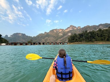 Ein Kind paddelt in einem Kajak auf einem ruhigen See, umgeben von Bergen und schwimmenden Hütten im Hintergrund.