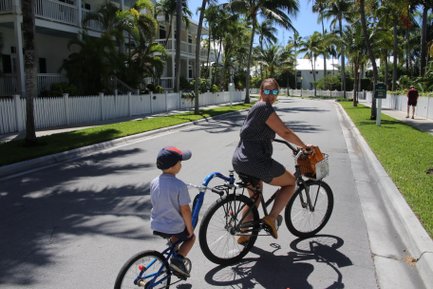 Eine Frau fährt mit einem Kind auf einem blauen Fahrrad auf einer sonnigen Straße, umgeben von Palmen und weißen Zäunen.