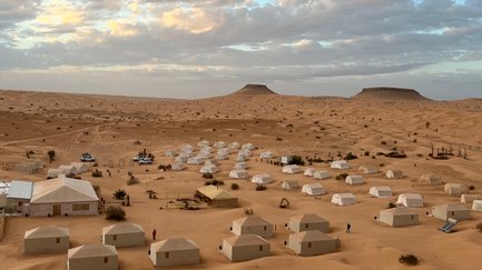 Eine weite Wüstenlandschaft mit zahlreichen Zelten, die in sanften Sanddünen angeordnet sind, unter einem bewölkten Himmel.