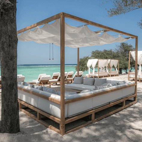 Ein elegantes Himmelbett aus Holz steht am Strand, umgeben von sanften Wellen und einem klaren blauen Himmel.