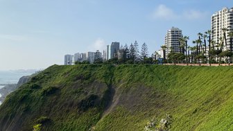 Eine grüne Klippe in Lima, Peru, mit modernen Gebäuden und Palmen im Hintergrund, unter einem klaren blauen Himmel.
