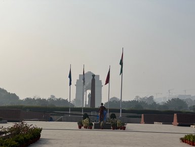 Ein Soldat steht in der Nähe des India Gate, umgeben von Flaggen und Pflanzen, während der Morgennebel die Szene umhüllt.