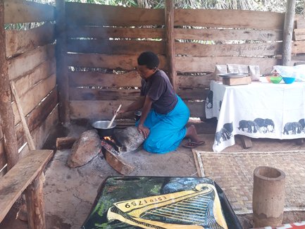 Eine Frau in blauer Kleidung kocht über einem offenen Feuer in einer einfachen Hütte, umgeben von Natur.