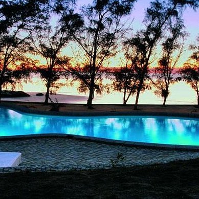 Ein glitzernder Pool in der Dämmerung, umgeben von hohen Bäumen und einem ruhigen Wasserblick auf den Sonnenuntergang.