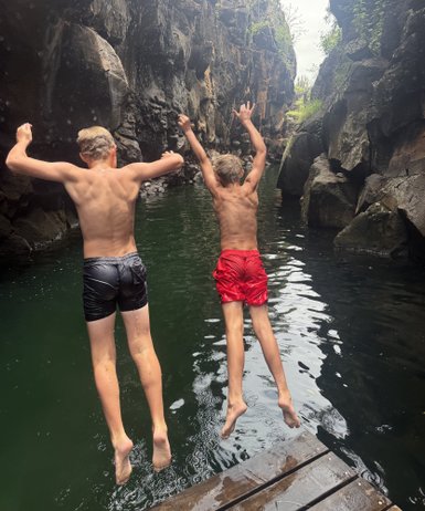 Zwei Jungen springen fröhlich in ein klares, grünes Wasser zwischen hohen, steilen Felsen.