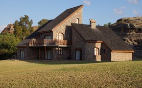 Ein modernes Steinhaus mit einem schrägen Dach steht in einer grünen Wiese, umgeben von sanften Hügeln und Bäumen.