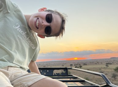 Ein Junge mit Sonnenbrille lächelt fröhlich aus einem Geländewagen, während die Sonne am Horizont untergeht.