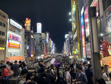 Die belebte Dotonbori-Straße in Osaka ist nachts erleuchtet, mit Menschenmengen, die am Fluss entlang spazieren.
