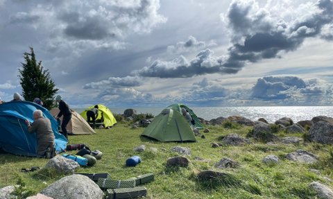 Eine Gruppe von Menschen baut Zelte am Ufer eines Sees auf, umgeben von großen Steinen und einer dramatischen Wolkenlandschaft.