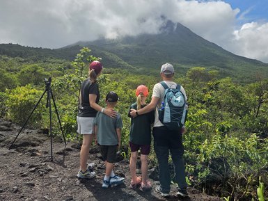 Eine Familie steht zusammen mit Blick auf den Vulkan Arenal, umgeben von üppiger Vegetation und Wolken.