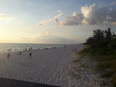 Ein belebter Strand bei Sonnenuntergang, mit Menschen, die am Wasser spazieren und im Sand entspannen.