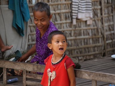 Ein fröhliches Kind mit kurzen Haaren trägt ein rotes Oberteil und schaut begeistert in die Kamera, während eine Frau im Hintergrund lächelt.
