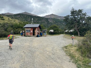 Ein Wanderer mit Rucksack geht auf einem Schotterweg zu einer kleinen roten Hütte in der Natur Patagoniens.