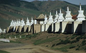 Eine Reihe von weißen Stupas erstreckt sich entlang einer grünen Wiese, umgeben von sanften Hügeln im Hintergrund.