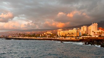 Die Küste von Puerto de la Cruz wird von sanften Wellen umspült, während die Sonne hinter den Wolken untergeht.