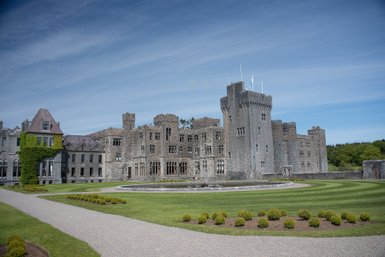 Ein beeindruckendes Schloss mit grauer Steinfassade und Türmen, umgeben von gepflegten Rasenflächen und Sträuchern.