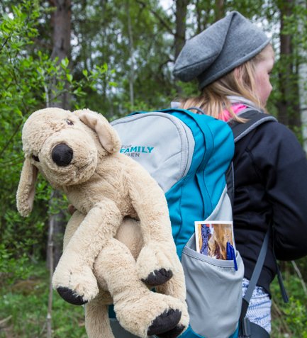 Ein Mädchen mit einem Rucksack, auf dem ein plüschiger Hund sitzt, steht in einer grünen Waldlandschaft.