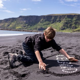 Ein Junge kniet am schwarzen Sandstrand und gestaltet mit Steinen eine kreative Form im Sand.