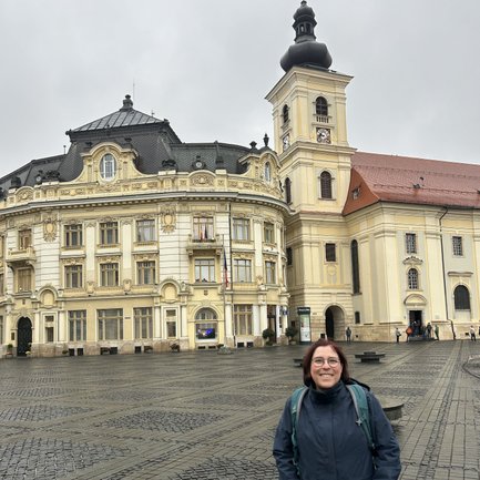 Eine Frau steht lächelnd auf einem Platz in Sibiu, umgeben von historischen Gebäuden und grauem Himmel.